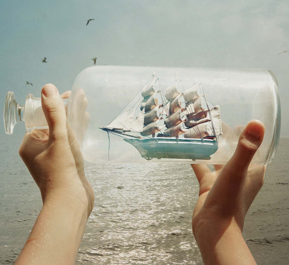 Small hands holding a detailed ship in a bottle against an brightly lit ocean backdrop